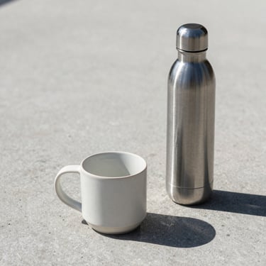 Minimalist ceramic mug and sleek metal water bottle on a concrete surface, sharp shadows, direct sunlight