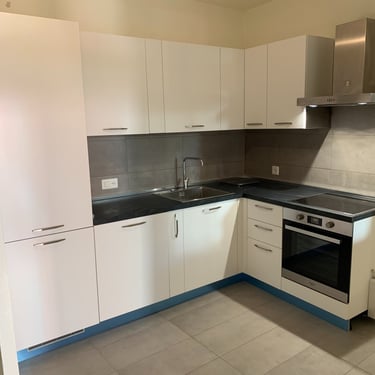 Cuisine en L blanche, plan de travail noir, crédence grise et électroménager intégré.