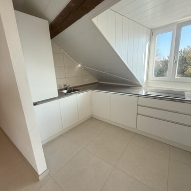 Cuisine sous combles blanche en L, plan de travail gris, rangements intégrés et fenêtre apportant lumière naturelle.