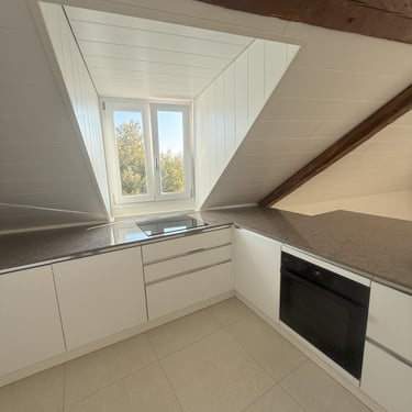 Cuisine sous combles blanche, plan de travail gris, plaque intégrée et fenêtre apportant une lumière naturelle.