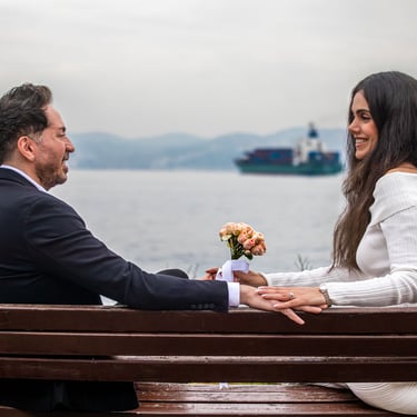 A smiling couple sits on a park bench by the water, celebrating an engagement with a floral bouquet.