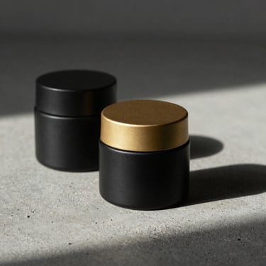 Minimalist ceramic skincare jars on a raw concrete surface, clean black label design, soft directional sunlight casting long elegant shadows.