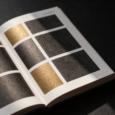 Close-up of an open editorial design book showing high-contrast grid layouts, heavy matte paper texture, dramatic shadows, warm studio light.