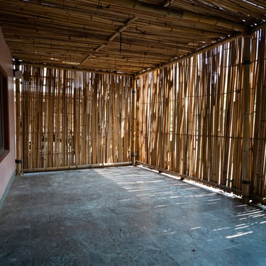 A spacious Lucknow home verandah shaded by floor-to-ceiling bamboo chicks, blocking the bright outdoor heat while keeping the tiled floor cool and private