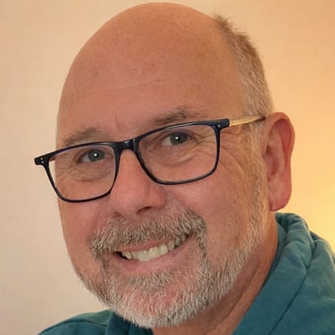 Smiling bald man with glasses and a grey beard wearing a blue shirt.