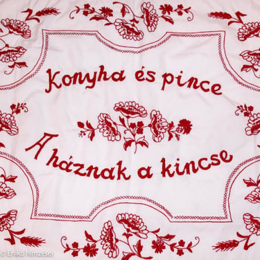 Piros fonallal készített falvédő szöveggel, virágos díszítéssel.