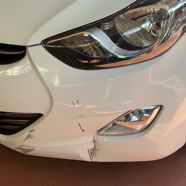Close-up of a white car's front bumper showing minor collision damage, paint scratches, and a dent