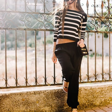 Mujer viste elegante viste suéter de rayas, pantalón de vestir negro y sandalias frente a un espejo moda internacional