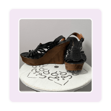 Pair of black strappy leather wedge sandals with high wood platform heels on a white stand.