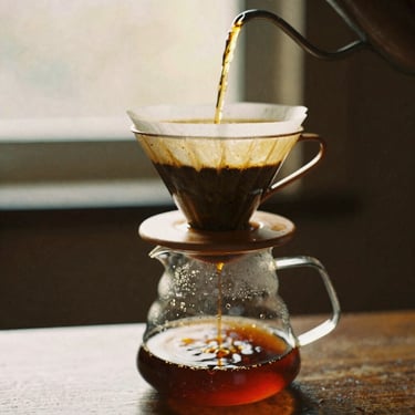 Pour-over coffee dripping slowly through a paper filter into a glass carafe, warm backlighting, rich amber tones.
