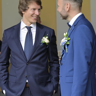 Zwei Hochzeitsgäste im Anzug stehen beim Sektempfang im Gespräch und lächeln sich an.