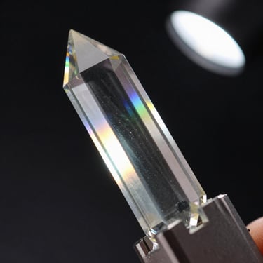 Macro close-up of a single Baccarat crystal prism held in a precision fixture, cool studio strobe from upper right, internal refraction visible as a sharp spectral band crossing the crystal's face, pure black background, no hands visible, no context — only the material and its optical behavior