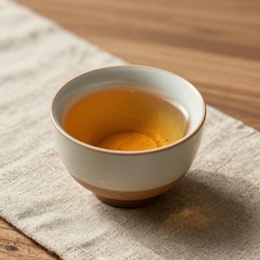 A single organic ceramic cup filled with warm tea sitting on a raw linen cloth, sunlit minimalist wooden table.