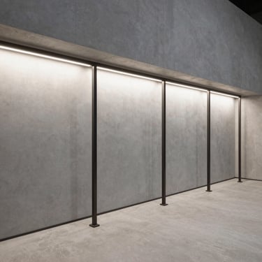 Fotografia de interior comercial minimalista. Piso de microcimento cinza claro, expositores lineares de metal escuro, iluminação zenital suave e uniforme. Linhas ortogonais limpas, sem pessoas.