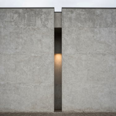 Fotografia em elevação plana de uma fachada minimalista de concreto cinza claro, abertura geométrica estreita revelando luz suave, sem pessoas.