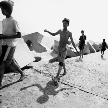 Group of boys walking barefoot on a sunlit harbour wall, expressive black-and-white seaside street scene.