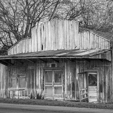 Abandoned and rundown building beside a rural road in Schwertner, Texas, with weathered wood, broken windows, and brush