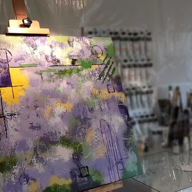 A purple, yellow, and green abstract painting on an easel with paints in the background