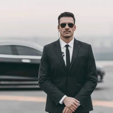 En livvakt står i kostym och solglasögon framför en svart bil.