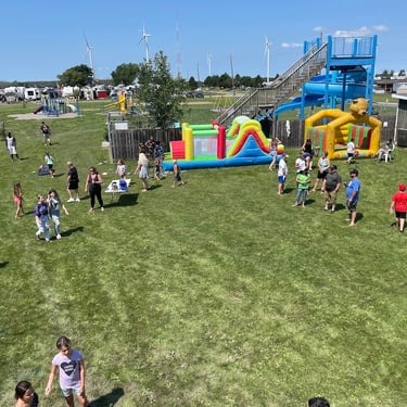 Families enjoy an outdoor summer festival at an RV park with inflatable bounce houses and a water slide.