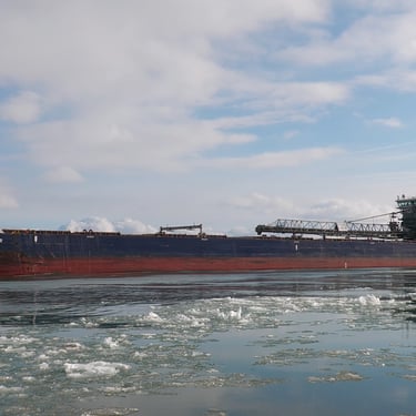 Freighter Algoma Bear from the front left side looking aft, on icy water with a plume of steam coming out of her funnel.