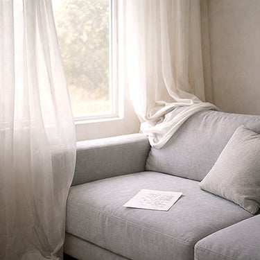 A softly lit couch by a window with sheer curtains and a single sheet of paper resting on the seat.