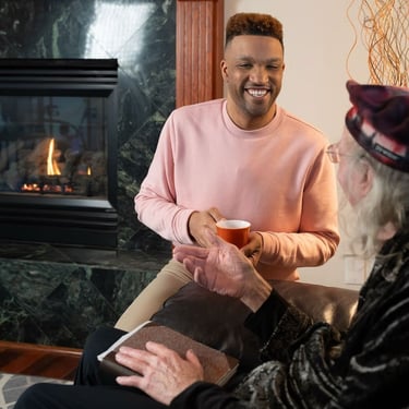 A smiling male caregiver holds a hot drink in a mug while conversing with an elderly man in front of a fireplace