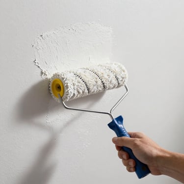 Close-up of a painter's roller applying white paint to a smooth wall, fresh coverage visible, clean drop cloth on floor, bright daylight