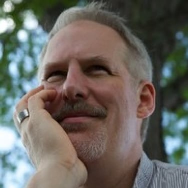 a smiling man with a mustache sitting outside