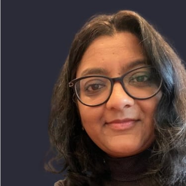 A woman with black rimmed glasses smiling slightly with wavy dark, shoulder length hair