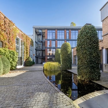Cour pavée avec bassin, bâtiments en briques couverts de lierre, arbustes taillés et façade de bureaux modernes.
