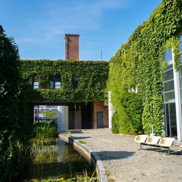 Cour pavée avec bassin, chaises et bâtiments couverts de lierre vert avec une cheminée en brique sous un ciel bleu.