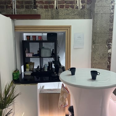 Espace pause avec mur en briques, frigo et étagère dans une alcôve, table haute blanche avec deux tasses noires.