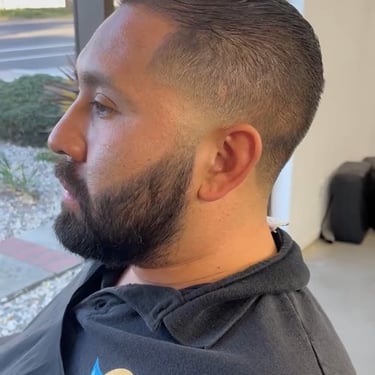 Side profile of a man with a neat beard and close fade haircut sitting near a window.