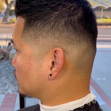 Person with a fade haircut seated inside near a window with trees outside.