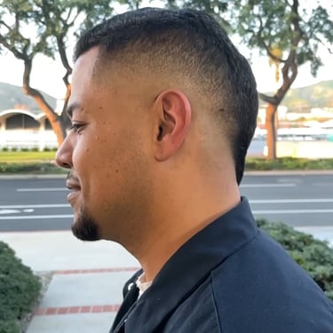 Side profile of a person outdoors with a fade haircut and a dark blue shirt, with trees and a road in the background.