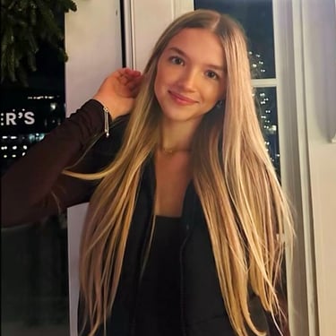 Emily smiling warmly indoors at night her long blonde hair as lights twinkle in the background