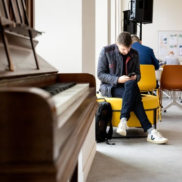 Une homme en veste noire assis sur un fauteuil jaune et regardant son smartphone