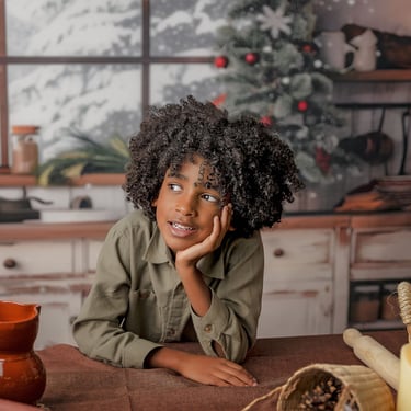 niño con pelo rizado apoyado en la mesa con ún árbol de navidad al fondo