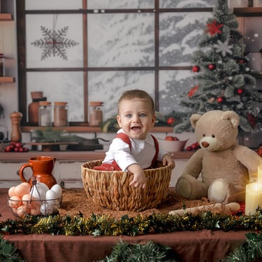 bebé sentado en una cesta al lado de un osito con ún árbol de navidad al fondo