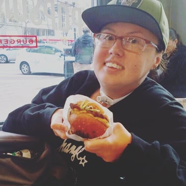 Photo of Stacey in a restaurant holding a chicken sandwich with a Raiders hat on.