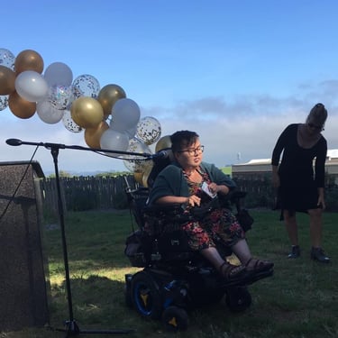 Photo of Stacey speaking at a graduation event, in front of microphone, with balloons in the background.