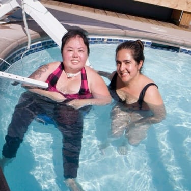 Photo of Stacey in a pool with friend. Stacey is not wearing glasses and has a little sunburn.