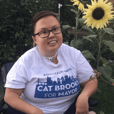 Picture of Stacey infront of a sunflower wearing a shirt reading "Cat Brooks for Mayor"