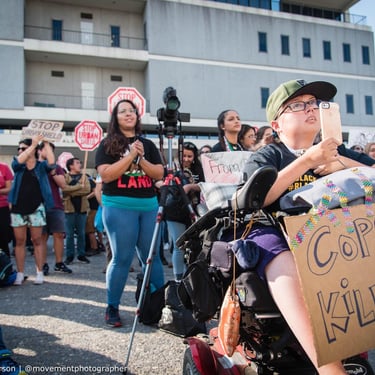 Photo of a protest, Stacey is in the foreground filming on her phone, holding a sign "Cops are Killing us"