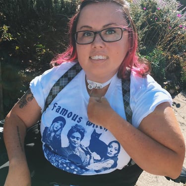 Photo of Stacey with suspenders, pink hair, tee shirt that says "Famous witches", with tattoos showing.