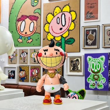 Contemporary urban vinyl art toy figure displayed in a gallery with colorful pop art paintings.