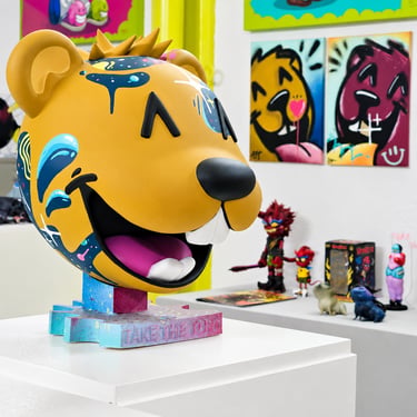 A large yellow designer art toy sculpture with colorful graffiti paint drips displayed in a modern art gallery.