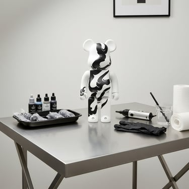 A black and white Bearbrick figure on a stainless steel table with tattoo ink bottles and supplies.