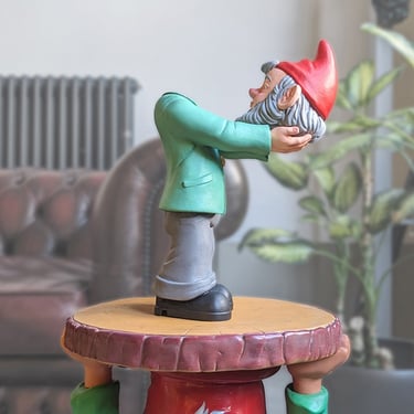 Quirky garden gnome figurine holding its own detached bearded head on a decorative pedestal.